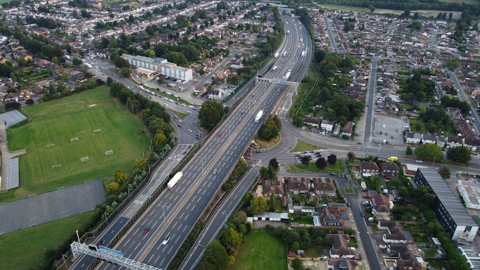 Aerial view of a multi-lane motorway with several trucks and vehicles in motion, crossing over urban residential and commercial areas within Abbey Wood and Thamesmead. The motorway features a central barrier and overhead signage equipment, with adjacent roads and roundabouts connecting to surrounding streets and neighborhoods. Nearby green spaces include sports fields and trees, while residential streets with detached and terraced houses are visible in the background. The scene is captured during daylight with clear weather, and the image showcases the logistical infrastructure relevant to house removals and furniture transport services between SE2 and SE28 areas, supported by Man with Van Crossness.
