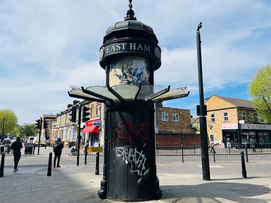 Photograph of a traditional black cylindrical town clock on a street corner in East Ham, featuring a domed top with ornamental detailing. The clock face is partially visible, surrounded by signage indicating 'East Ham.' Nearby, there are pedestrians walking on the pavement, with some standing near a small shop with a red and white awning. Several residential buildings and a nail salon are visible in the background, along with a clear blue sky and a few trees. The scene depicts a typical urban environment suitable for house removals and relocation services, with potential for loading and unloading furniture or boxes from nearby vehicles, as managed by companies like Man with Van Crossness during a home relocation process.
