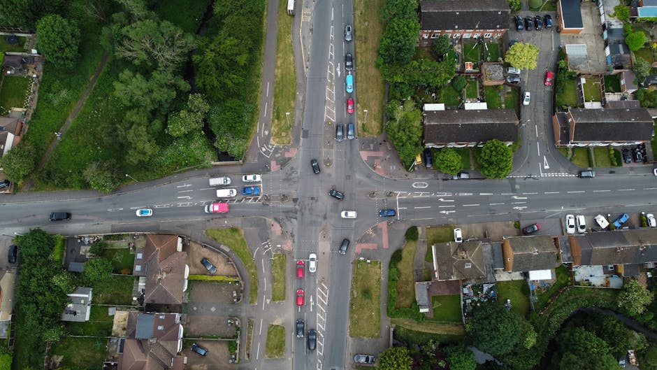 Aerial view of a multi-lane motorway with several trucks and vehicles in motion, crossing over urban residential and commercial areas within Abbey Wood and Thamesmead. The motorway features a central barrier and overhead signage equipment, with adjacent roads and roundabouts connecting to surrounding streets and neighborhoods. Nearby green spaces include sports fields and trees, while residential streets with detached and terraced houses are visible in the background. The scene is captured during daylight with clear weather, and the image showcases the logistical infrastructure relevant to house removals and furniture transport services between SE2 and SE28 areas, supported by Man with Van Crossness.