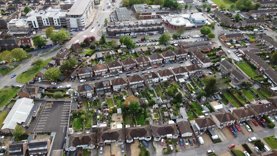 An aerial view of a residential neighbourhood showing rows of semi-detached houses with small gardens and driveways, parked cars along the streets, and some houses with solar panels on their roofs. In