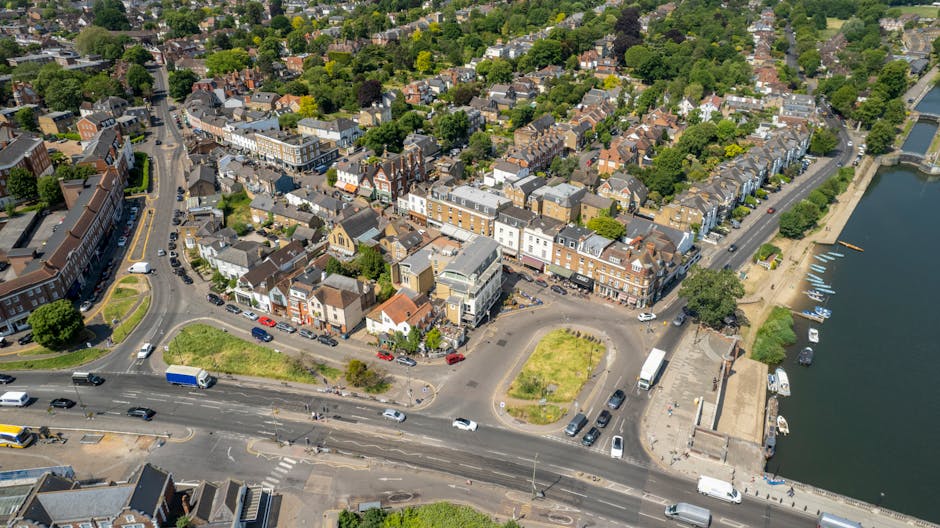 An aerial view of a residential neighbourhood showing rows of semi-detached houses with small gardens and driveways, parked cars along the streets, and some houses with solar panels on their roofs. In