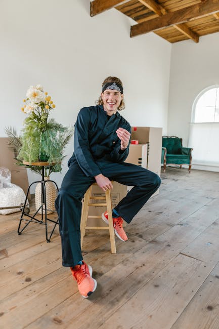 A young woman with light skin, wearing a dark blue work uniform, a black bandana, and bright orange sneakers, is seated on a small wooden stool inside a bright, airy room. She is smiling and gesturing with her right hand. Her hair is pulled back, and she appears relaxed and cheerful. The room has a high, angled wooden ceiling and large arched window allowing natural light to flood in, illuminating the wooden floorboards. To her left, there is a small round table with a green metal frame, holding a clear glass vase filled with white and yellow flowers, and a small potted plant beside it. In the background, there are cardboard boxes stacked near a cream-colored wall with a large, rounded window, and a green upholstered armchair near the wall. The scene suggests a home environment during a furniture or house removal process, with [COMPANY_NAME] providing the logistics of packing, loading, or transport involved in house removals, as part of a professional move at [PAGE_TITLE].