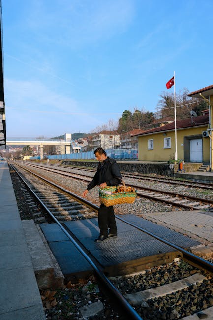 A man wearing a dark jacket and black trousers stands on a railway crossing operated by Man with Van Crossness, holding a wicker basket filled with various items, possibly packing materials or personal belongings, as part of a home relocation process. He is positioned on a safety platform covering the railway tracks, which are laid with wooden sleepers and metal rails, and appears to be preparing to cross or load items onto a nearby vehicle. In the background, there is a small pale yellow building with white window frames and a red Turkish flag on a tall pole, indicating an outdoor location likely near residential or industrial premises. The scene is set during daylight with clear blue skies and some trees visible behind the buildings, emphasizing the outdoor environment involved in the furniture transport or packing and moving activities associated with house removals. The image underscores aspects of moving logistics, including careful handling of possessions during loading or unloading near the railway infrastructure.