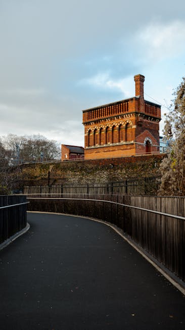 A young woman with light skin, wearing a dark blue work uniform, a black bandana, and bright orange sneakers, is seated on a small wooden stool inside a bright, airy room. She is smiling and gesturing with her right hand. Her hair is pulled back, and she appears relaxed and cheerful. The room has a high, angled wooden ceiling and large arched window allowing natural light to flood in, illuminating the wooden floorboards. To her left, there is a small round table with a green metal frame, holding a clear glass vase filled with white and yellow flowers, and a small potted plant beside it. In the background, there are cardboard boxes stacked near a cream-colored wall with a large, rounded window, and a green upholstered armchair near the wall. The scene suggests a home environment during a furniture or house removal process, with [COMPANY_NAME] providing the logistics of packing, loading, or transport involved in house removals, as part of a professional move at [PAGE_TITLE].