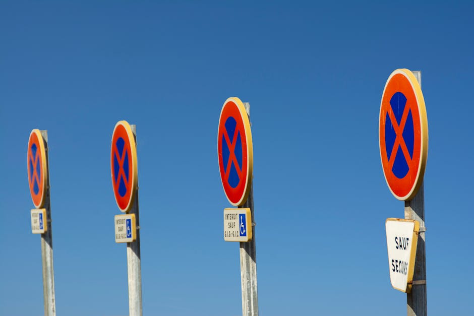 Four tall, metal parking restriction signs with blue and orange no-parking symbols are displayed against a clear blue sky. The signs are positioned in a row on a paved surface, with each sign featuring an additional small white rectangle indicating parking allowances, including one with a wheelchair symbol for accessible parking. The signs are part of a street scene adjacent to a residential property on Crossness Road. The image captures the signs from a slightly low angle, emphasizing their height and uniformity. Some of the signs include white warning labels with black text, indicating restrictions or notices, such as 'SAUF', suggesting specific exemptions. The environment suggests an urban or suburban area, with the signage potentially impacting parking arrangements for home access or relocating vehicles during house moving or furniture transport processes, as managed by Man with Van Crossness, specialists in removals and home relocation services.