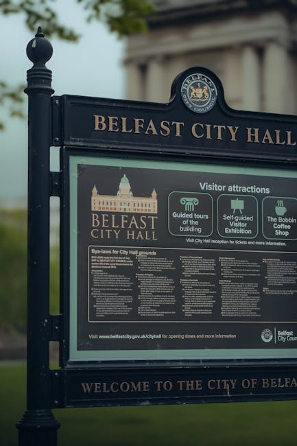 A large, free-standing information board positioned outdoors next to Belfast City Hall, with a dark blue metal frame and decorative finial at the top. The board displays details about Belfast City Hall, including visitor attractions such as guided tours, self-guided exhibitions, and the Bobbin Coffee Shop, with icons representing each attraction. Below the main information, there are official bye-laws for the city hall grounds presented in small print. The background features blurred trees and a cloudy sky, indicating a park or outdoor civic area. The setting suggests a location where visitors might park their vehicles or begin their house relocation or furniture transport process, with the Belfast City Hall sign subtly supporting the context of local moving regulations and services provided by companies like Man with Van Crossness.