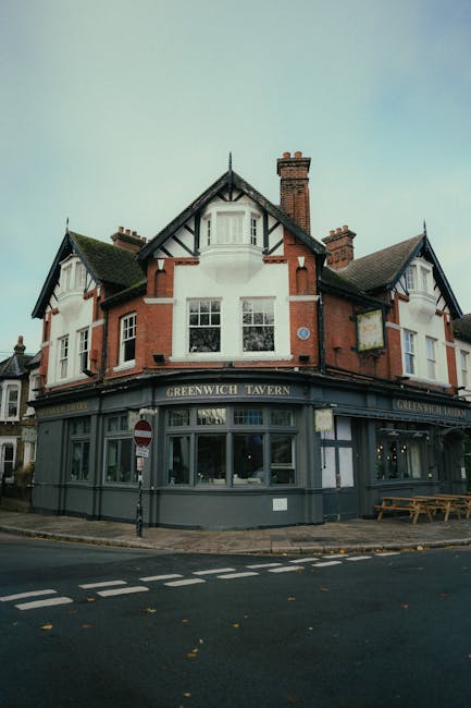 A three-story, red-brick building with white window frames and decorative black timbering on the upper floor, housing the Greenwich Tavern pub. The ground floor features large glass windows and a sign displaying the pub's name, with an outdoor seating area comprising wooden benches and tables situated on the pavement in front of the entrance. The building is located at a street corner, with a black and white 'no entry' traffic sign and a pole supporting street signs visible near the sidewalk. The scene appears to be during daytime with partly cloudy weather, and no moving activity is visible. This setting represents a typical exterior view relevant to house and pub relocations or transportation preparations, as involved in effective packing and moving logistics, supported by Man with Van Crossness' services.
