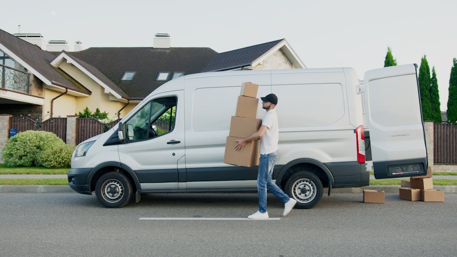 A young man with long, wavy blond hair, wearing a dark bandana and a navy blue shirt, is sitting in the driver's seat of a white moving van during daytime. He is smiling at the camera and giving a thu