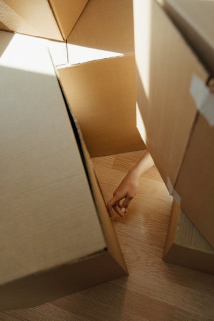 A human hand reaching into an open cardboard box during a home relocation process, with the box positioned on a light wooden floor. Surrounding the opening are several other cardboard boxes stacked and arranged, some sealed with packing tape and others partially open, indicating packing and moving activities. Natural light illuminates the scene, suggesting an indoor environment, typically seen during furniture transport and packing for house removals by Man with Van Crossness. The image captures the careful handling involved in loading or unpacking bulky items without damage, integral to professional removals services like those provided in Thamesmead flats.