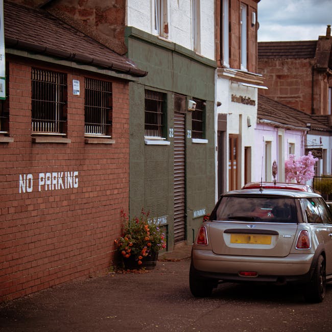 A silver hatchback vehicle parked on a narrow pavement in front of a row of buildings with varied facades, including a red brick structure with barred windows and a green-painted commercial unit with a roller shutter door, indicating a residential or small business area. To the left, a white painted sign on the brick wall reads 'NO PARKING,' and a flower pot with colorful blooms is placed below the sign. The scene is set during daylight with some clouds visible in the sky, and the vehicle appears to be in the process of a home relocation or furniture transport, possibly managed by Man with Van Crossness, as part of a removal or moving service. The setup suggests that the vehicle may be loading or unloading household items close to the doorway of the nearby property, with the surrounding environment emphasizing urban residential relocation logistics.