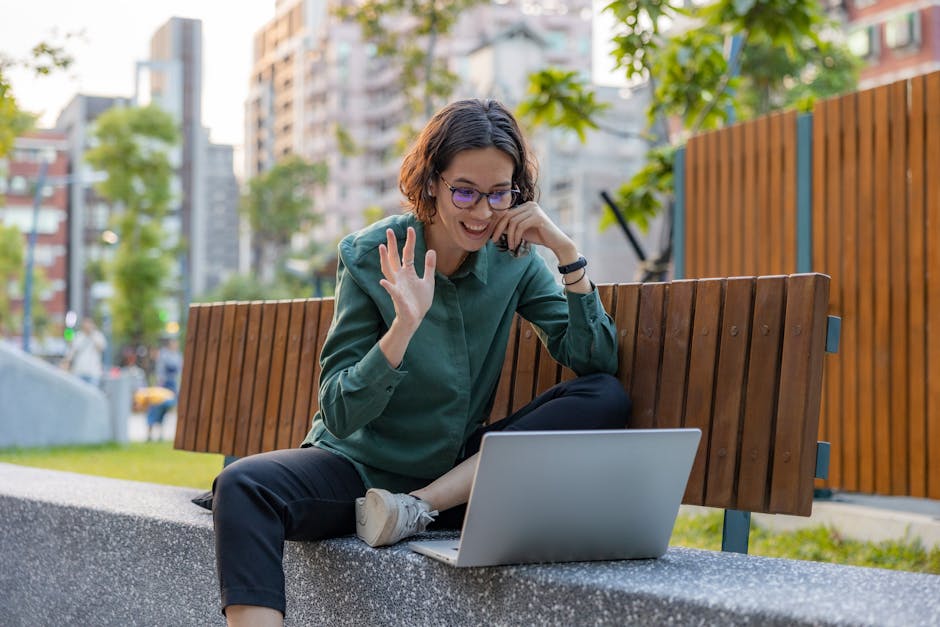 A young woman with shoulder-length brown hair, wearing glasses, a green shirt, and dark trousers, is sitting on a concrete ledge in an outdoor urban setting. She is engaged in a video call on her laptop, which rests on her lap, and is smiling while waving with her right hand. Behind her, there is a wooden fence, some green trees, and tall residential buildings. The scene is well-lit with natural daylight, indicating it is daytime, and the woman appears relaxed and happy. This image relates to home relocation and moving logistics, as it captures a moment of communication possibly connected to planning or coordinating a move, and represents the friendly, professional service offered by Man with Van Crossness in house removals.