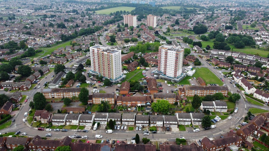 An aerial view of a residential area in Thamesmead, featuring an arrangement of houses, low-rise apartment buildings, and high-rise flats with red and white facades. The foreground shows a curved street lined with parked cars and detached houses with small gardens in front. Behind this, there are larger apartment blocks located within a green space, with parking areas adjacent to them. The background extends to a suburban landscape with more houses, roads, and open green areas, all under a cloudy sky. This image captures the layout of a typical suburban neighbourhood, illustrating the mix of housing types and the transportation infrastructure, relevant for visual context related to house removals and moving logistics in Thamesmead. Man with Van Crossness provides professional removals services in this area, supporting residents with residential relocations, furniture transport, and packing and moving processes, as seen in the context of home relocation activities.