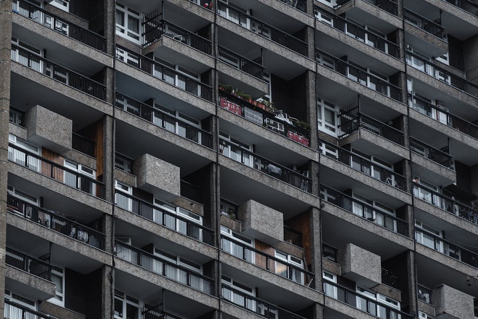 Close-up view of a high-rise residential building with multiple balconies, each featuring metal railings and sliding glass doors, some open and revealing interior spaces with curtains and plants. The building's exterior is constructed of grey concrete with textured surfaces, and several balconies display personal items such as potted plants, outdoor furniture, and clothing hanging to dry. The image captures the uniform pattern of the balconies across the facade, with natural daylight illuminating the scene, indicating daytime. This setting exemplifies a typical urban apartment block context relevant to home relocation and moving services, illustrating the complexity of accessing various levels for furniture transport and packing during the moving process. Man with Van Crossness may facilitate the logistics of moving household items from such multi-story properties, ensuring careful handling of furniture and belongings during the loading process into cachet-protected vans, visible at the ground level, ready for transport. The image highlights the importance of house removals experts managing loading and unloading in multi-floor residential environments.