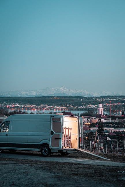 A grey moving van parked on an outdoor construction or staging area during dusk, with its rear doors wide open revealing a wooden furniture piece wrapped in protective plastic and partially inside the